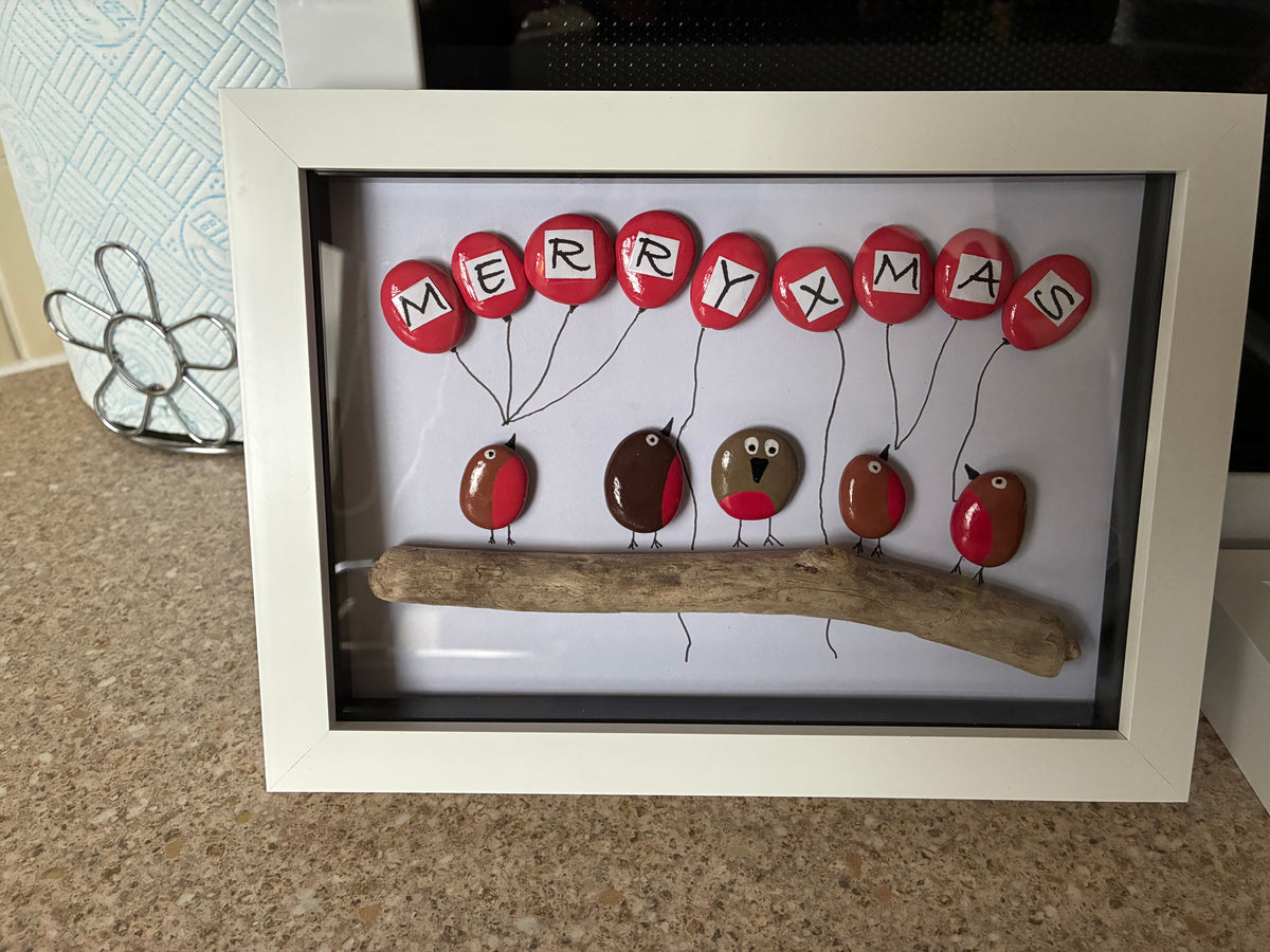 Five robin pebbles on driftwood holding red balloon pebbles spelling Merry Xmas in a white frame