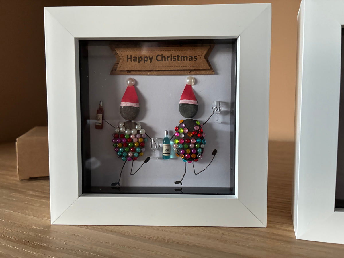 Two pebble friends in Santa hats decorated with gems and pearls sharing drinks in a white frame