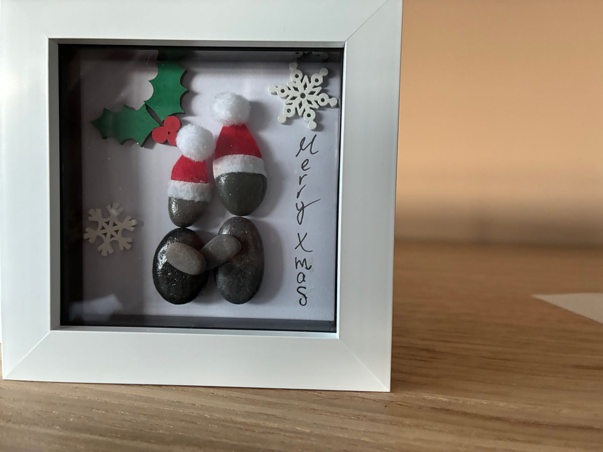 Two pebble people in Santa hats sharing a festive hug with holly and snowflakes in a white frame
