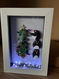Penguin pebbles decorating a sea glass Christmas tree with LED lights in a white frame