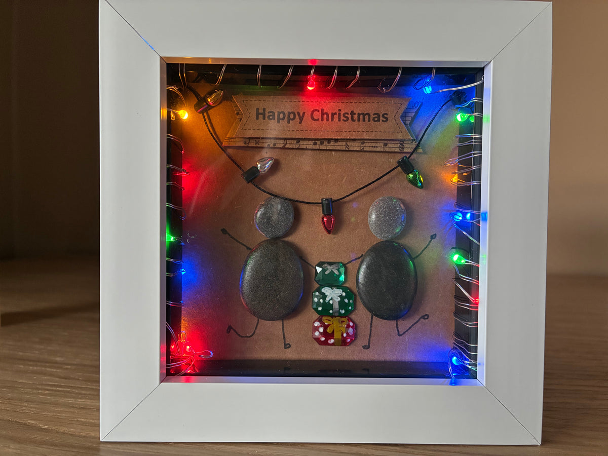 Two pebble friends sharing Christmas gifts with multicolour LED lights in a white frame