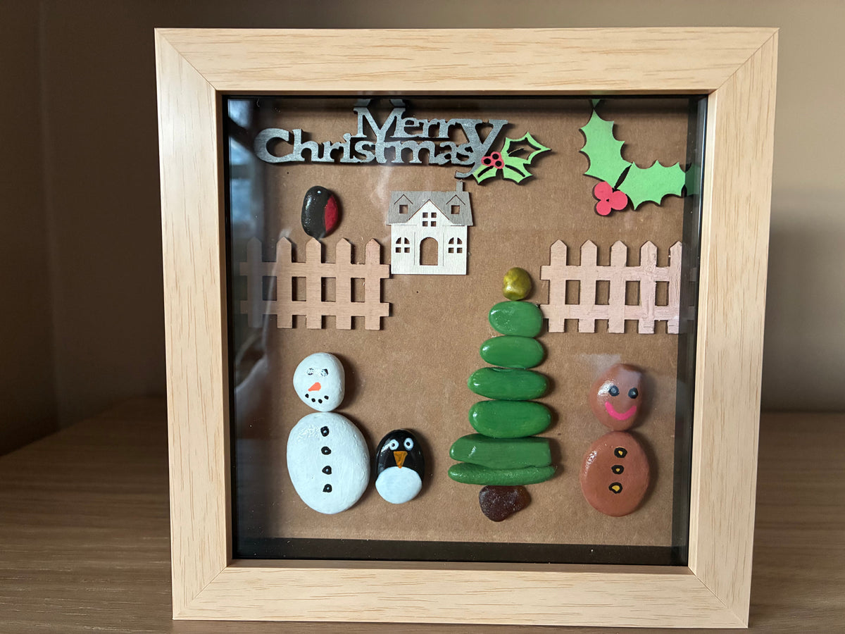 Festive Christmas pebble art scene with snowman, penguin, Christmas tree, gingerbread man and robin in a wooden frame