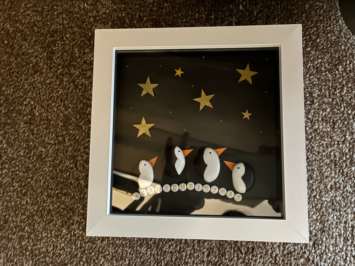 Handmade pebble art frame featuring three penguin figures under gold stars with Merry Christmas pebble lettering