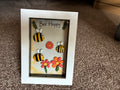 Bee Happy pebble art shadow box frame with three hand-painted bee pebbles, coral and orange pebble daisies, and wooden leaf embellishments on a yellow watercolour background, in a white frame