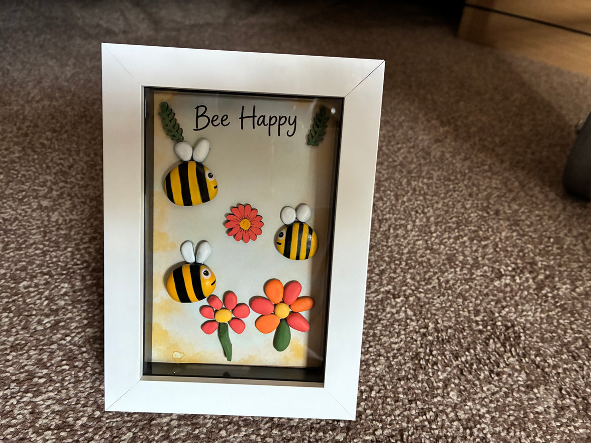 Bee Happy pebble art shadow box frame with three hand-painted bee pebbles, coral and orange pebble daisies, and wooden leaf embellishments on a yellow watercolour background, in a white frame