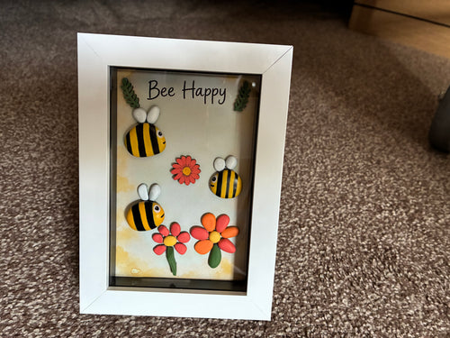 Bee Happy pebble art shadow box frame with three hand-painted bee pebbles, coral and orange pebble daisies, and wooden leaf embellishments on a yellow watercolour background, in a white frame