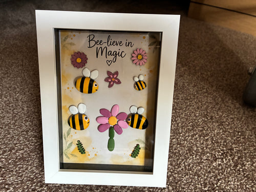 Bee-lieve in Magic pebble art shadow box frame with four hand-painted bee pebbles, a pink pebble daisy, and wooden flower embellishments on a watercolour botanical background, in a white frame