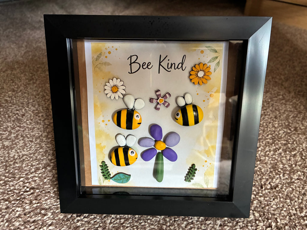 Bee Kind pebble art shadow box frame with three hand-painted bee pebbles, a purple pebble flower, and wooden flower embellishments on a yellow watercolour background