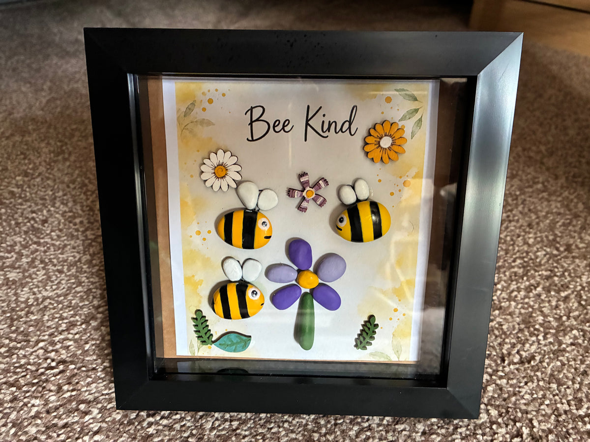 Bee Kind pebble art shadow box frame with three hand-painted bee pebbles, a purple pebble flower, and wooden flower embellishments on a yellow watercolour background