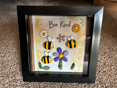 Bee Kind pebble art shadow box frame with three hand-painted bee pebbles, a purple pebble flower, and wooden flower embellishments on a yellow watercolour background